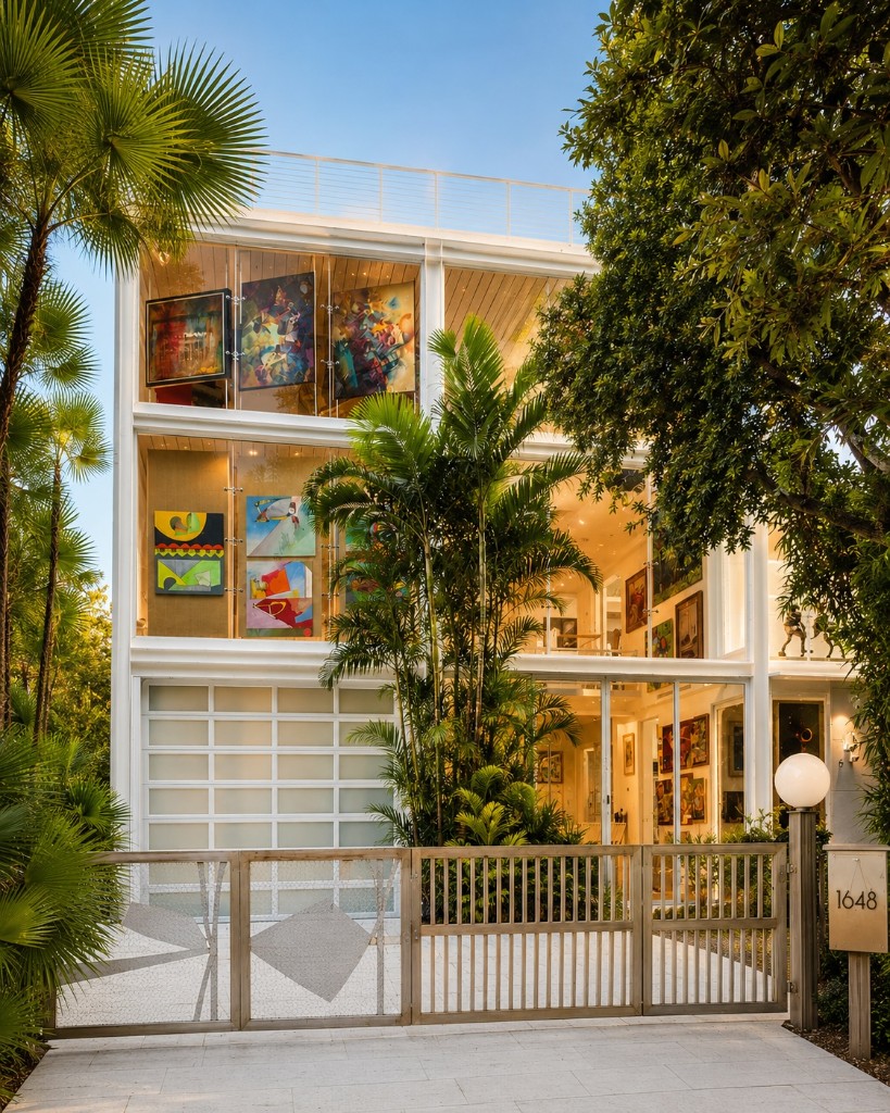 Vertical exterior photo of a modern tropical house in St. Petersburg, Florida, featuring a glass-front façade, lush tropical landscaping, and colorful artwork visible from outside.